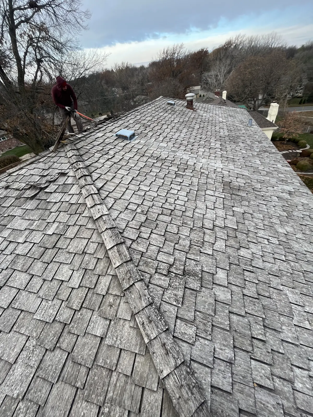 Weathered wood shake roof before replacement, with a roofer up at the ridge